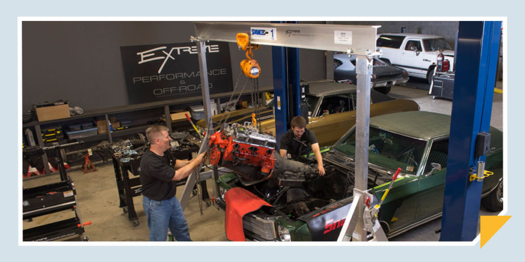 Two mechanics using a spanco crane to help remove an engine from a green car.